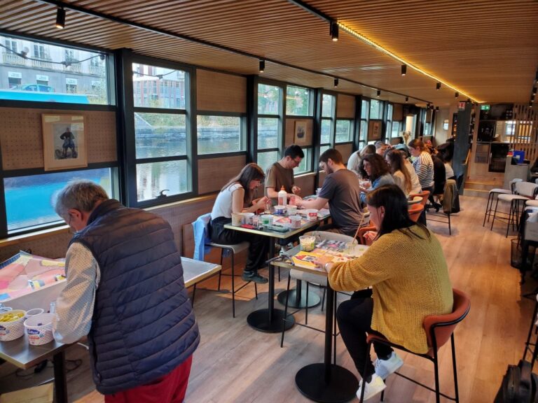 Tout le monde est attablé et concentré sur son travail à réaliser en mosaïque. C'est sur une péniche et sont sur des tables hautes.