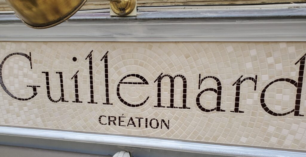 Vue de la mosaïque avec le nom Guillemard Création, les couleurs choisies sont le marron pour l'écriture et des tons blancs cassés et beiges pour l'entourage en mosaïque