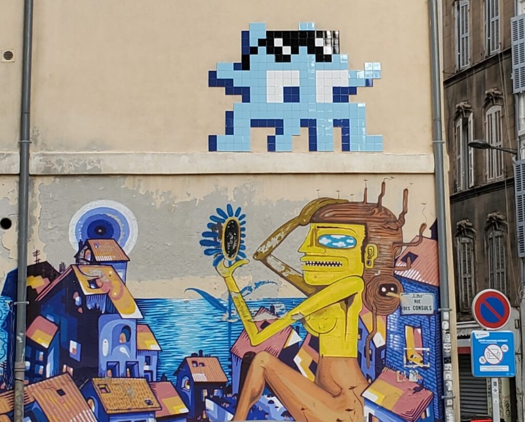 Personnage Space Invader réalisé en mosaïque de pixels aux couleurs bleu ciel. Ce personnage est au-dessus d'une fresque réalisée à la bombe représentant une femme et un homme