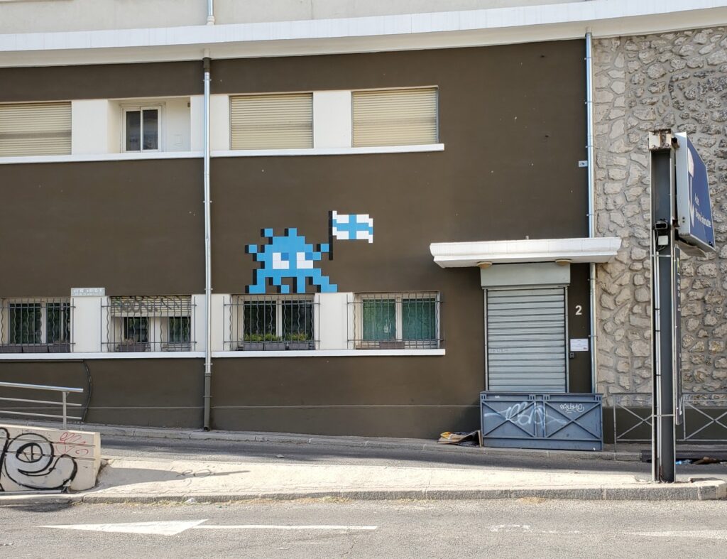 Sur cette photo d'un parking, il y a un personnage bleu et blanc réalisé en mosaïque de pixels avec un drapeau grec