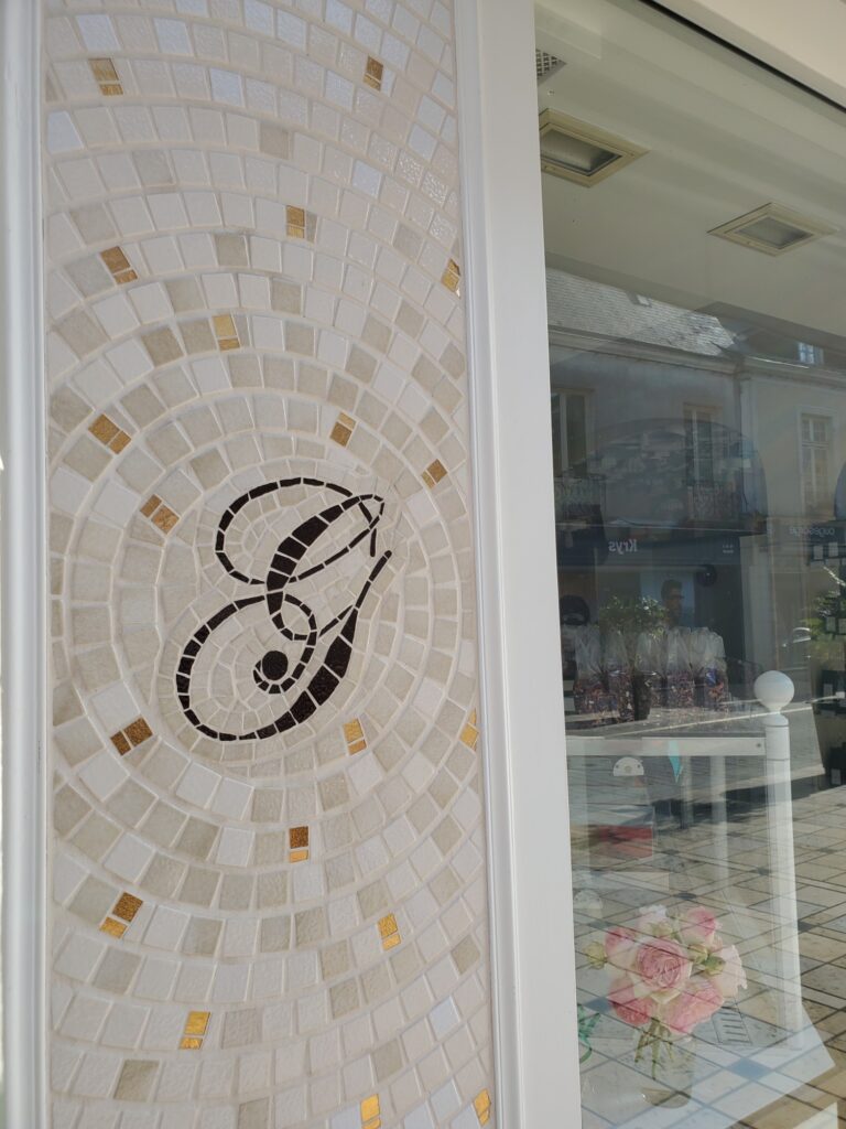 panneau de gauche d'une enseigne de pâtisserie. Il s'agit d'un G en mosaïque marron avec des cercles et ondulations en mosaïque beige, blance, dorée. Réalisée par Céline Loury