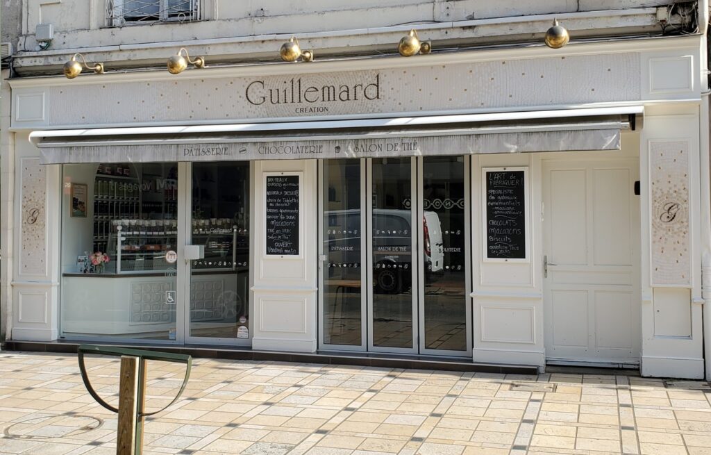 Enseigne d'une pâtisserie en mosaïque. Sur la photo, il y a les pavés, puis une devanture peinte en blanc cassé. Sur le panneau du haut, il y a le mot Guillemard création d'écrit en mosaïque en marron - couleur cacao. Ce mot est entouré d'ondulations en mosaïque en dégradés de tons clairs, beiges, dorés. Sur les côtés, deux panneaux avec le G de l'identitié visuelle de la pâtisserie en mosaïque.