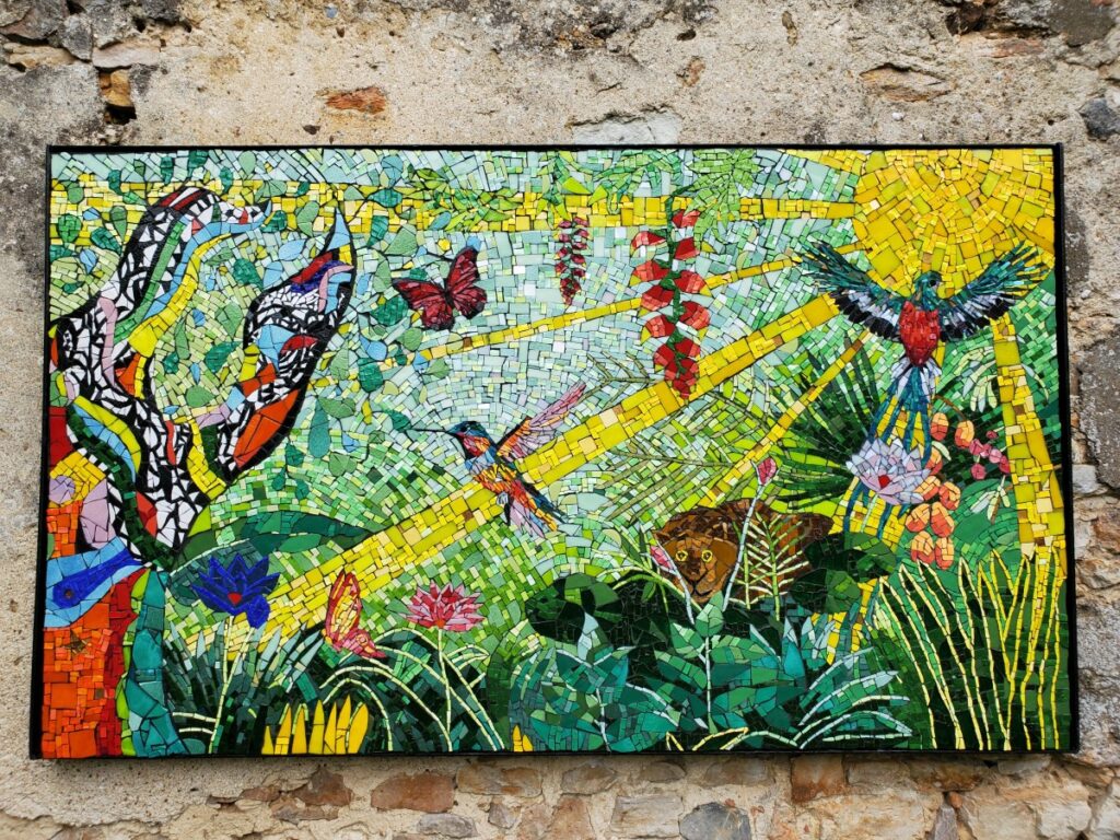 Fresque en mosaïque, sur ce décor, il y a l'arbre coloré de Niki de st Phalle à gauche. Puis un colibir en mosaïque, un quetzal en mosaïque, une inspiration du Douanier Rousseau, et les rayons du soleil pour avoir le soleil toute l'année dans le jardin. Fresque réalisée à quatre mains avec Céline Loury et Fanny Guillo