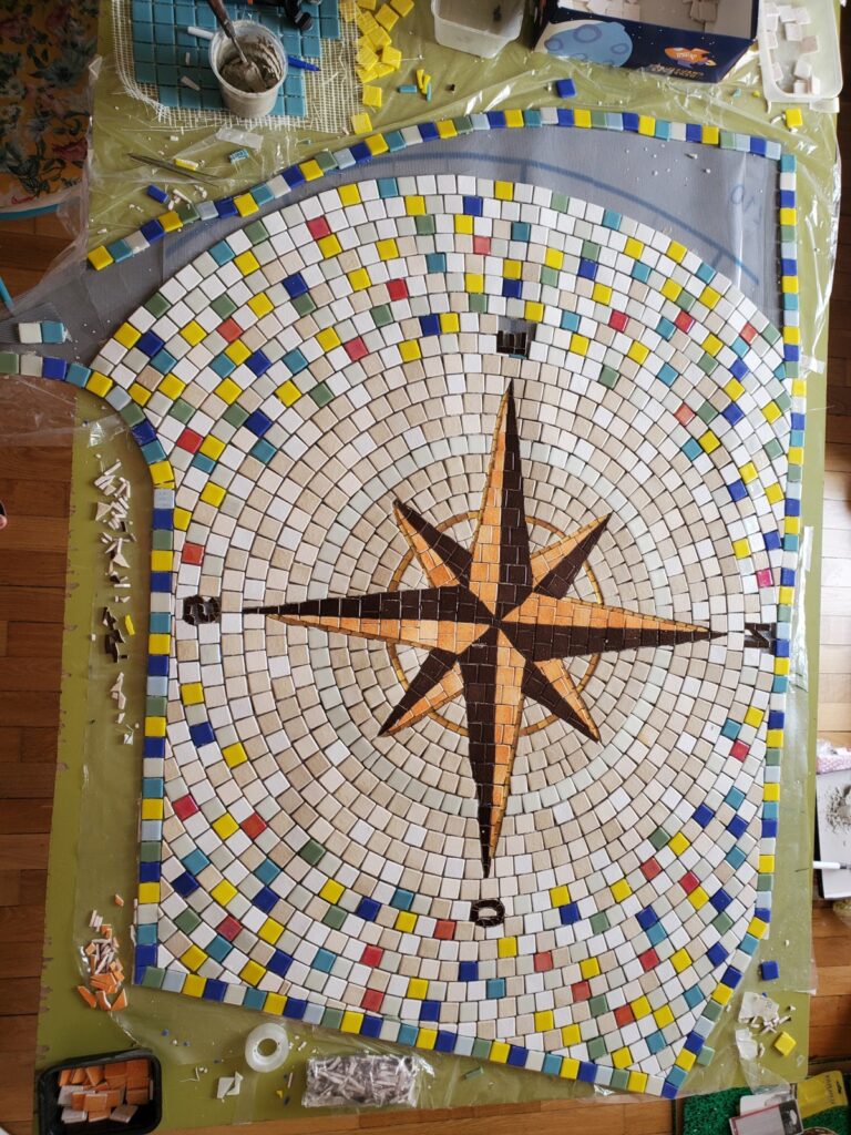 rose des vents de couleur orange et marron, avec un fond blanc et beige et quelques couleurs, une création en mosaïque et émaux de Briare par Céline Loury